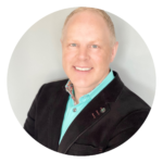 Blond man in black jacket and teal shirt smiles, professional headshot