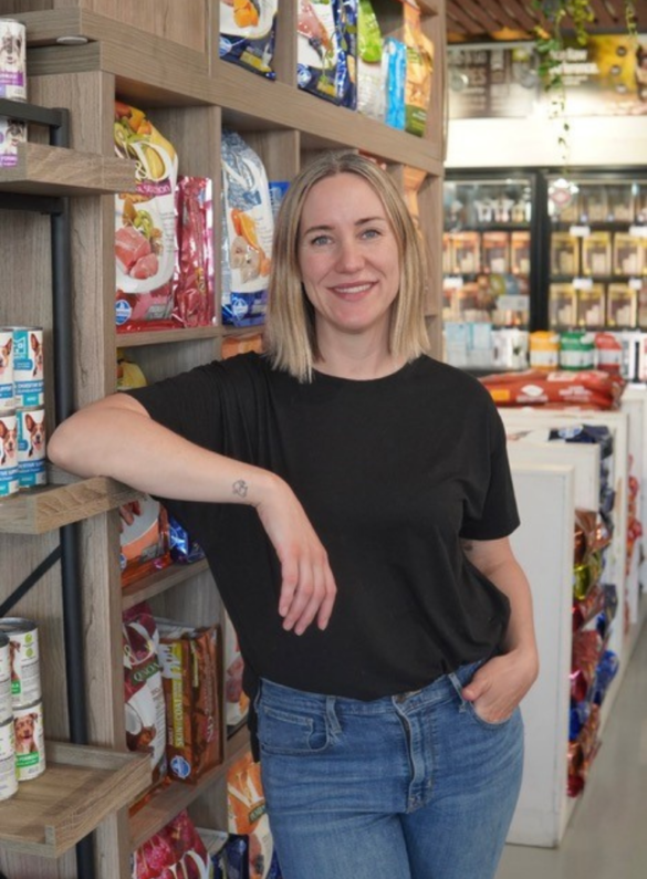 Shopkeeper leans on shelves stocked with pet food, smiling confidently.