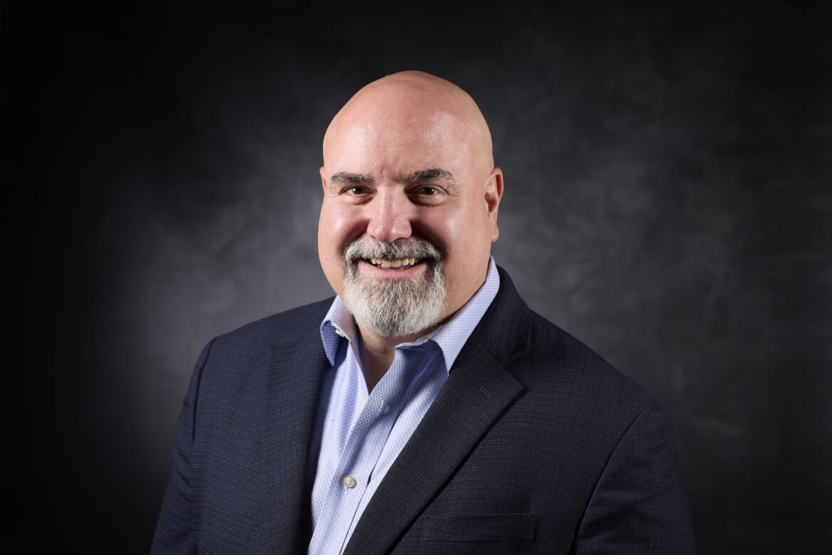 Bald man with a goatee smiling in a dark blazer and light blue shirt