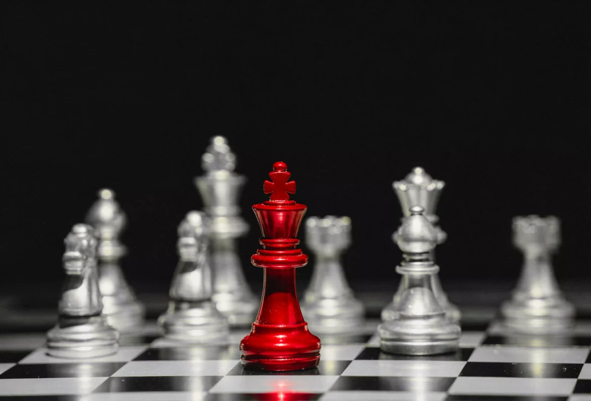 Red king chess piece stands tall centre with silver pieces blurred behind on chessboard