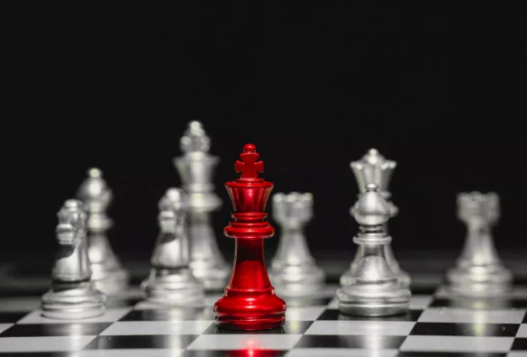 Red king chess piece stands tall centre with silver pieces blurred behind on chessboard