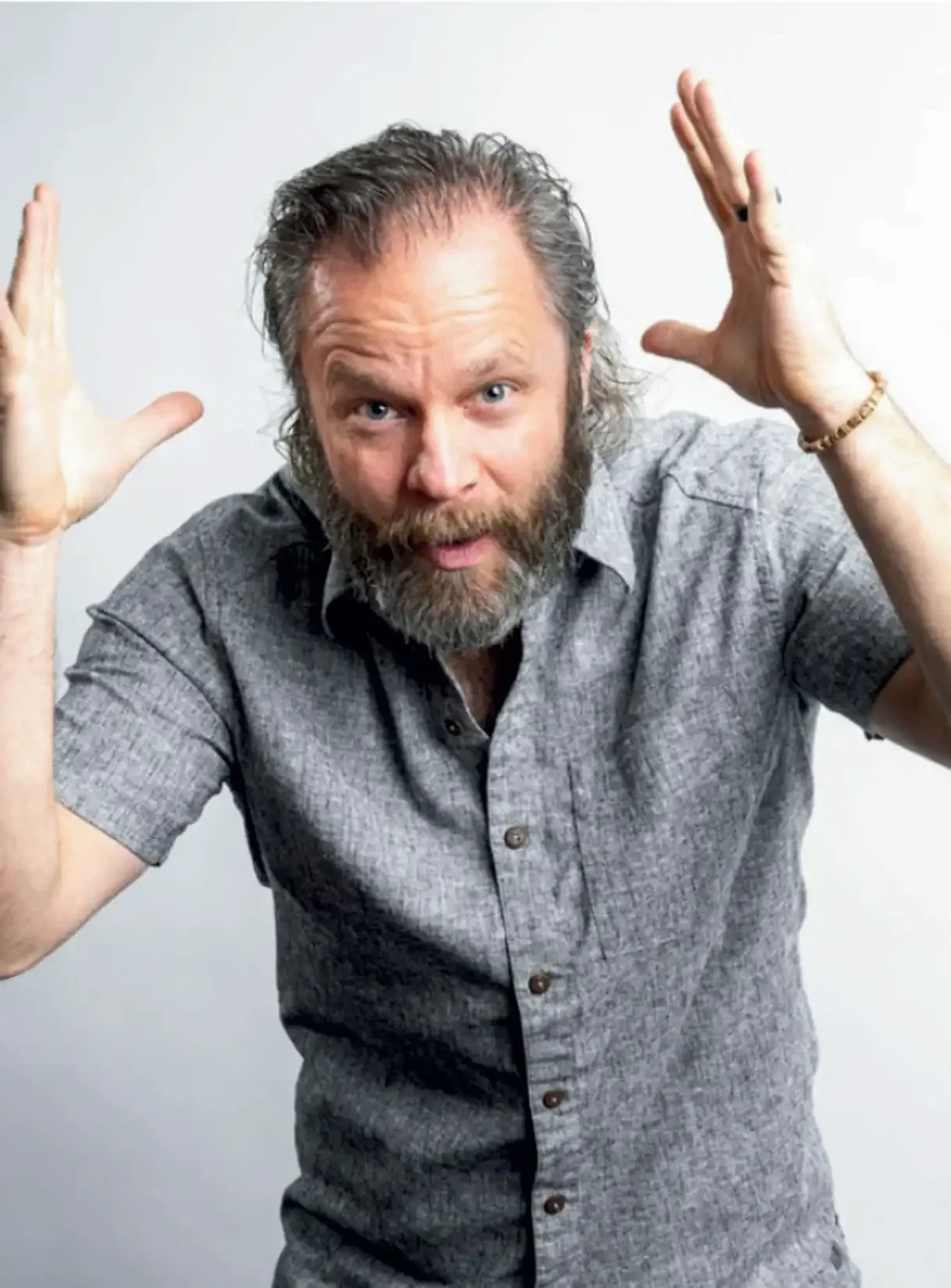 Man with grey hair and beard in grey shirt with hands raised