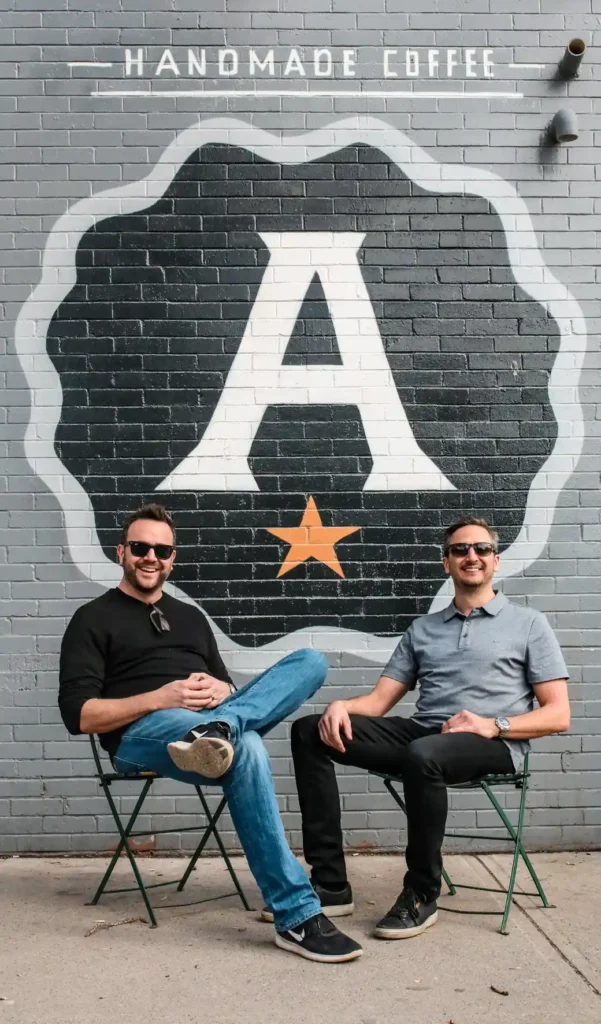 Two men sitting in front of a brick wall mural with a large A and "Handmade Coffee" text.