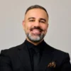 Man with beard wearing a black suit and tie with a patterned pocket square, smiling.