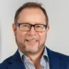 Man with beard and glasses wearing a blue blazer and checkered shirt smiling