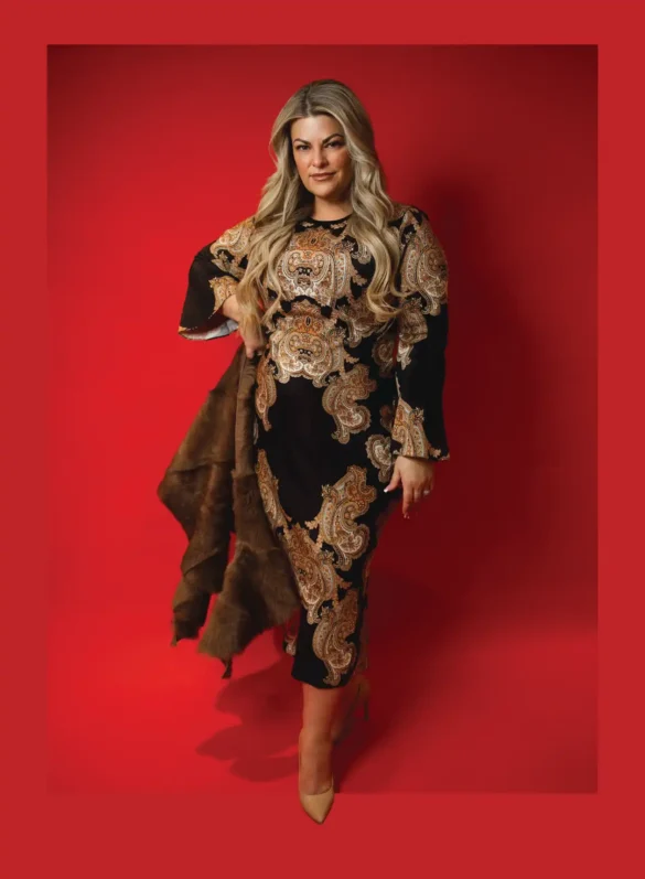 Woman in black paisley dress and fur sash stands against red background