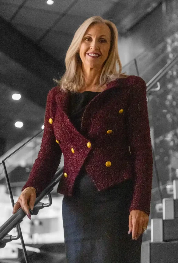 Blonde woman in a burgundy blazer with gold buttons smiles by a staircase.