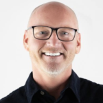 Bald man with glasses and a beard smiles, wearing a dark shirt.