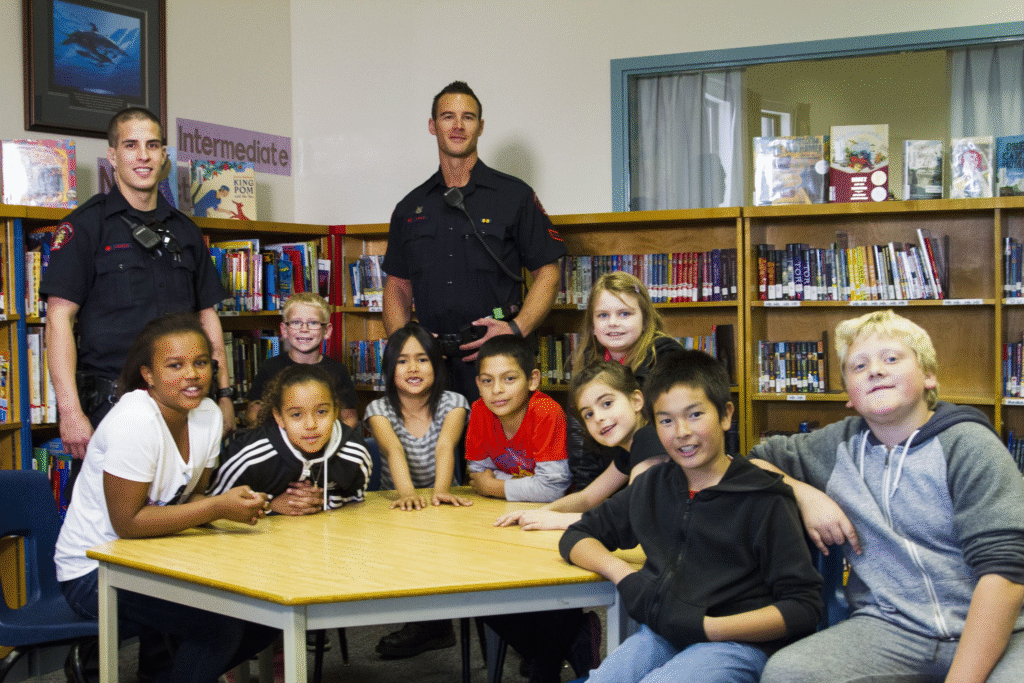 Firefighters pose with students in a library with shelves of books.