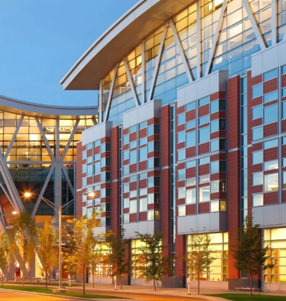 Modern building with brick, glass and metal facade illuminated at dusk walkway with trees and lampposts