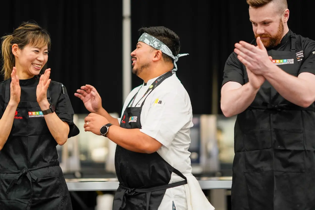 Three chefs in black aprons clap enthusiastically on stage.