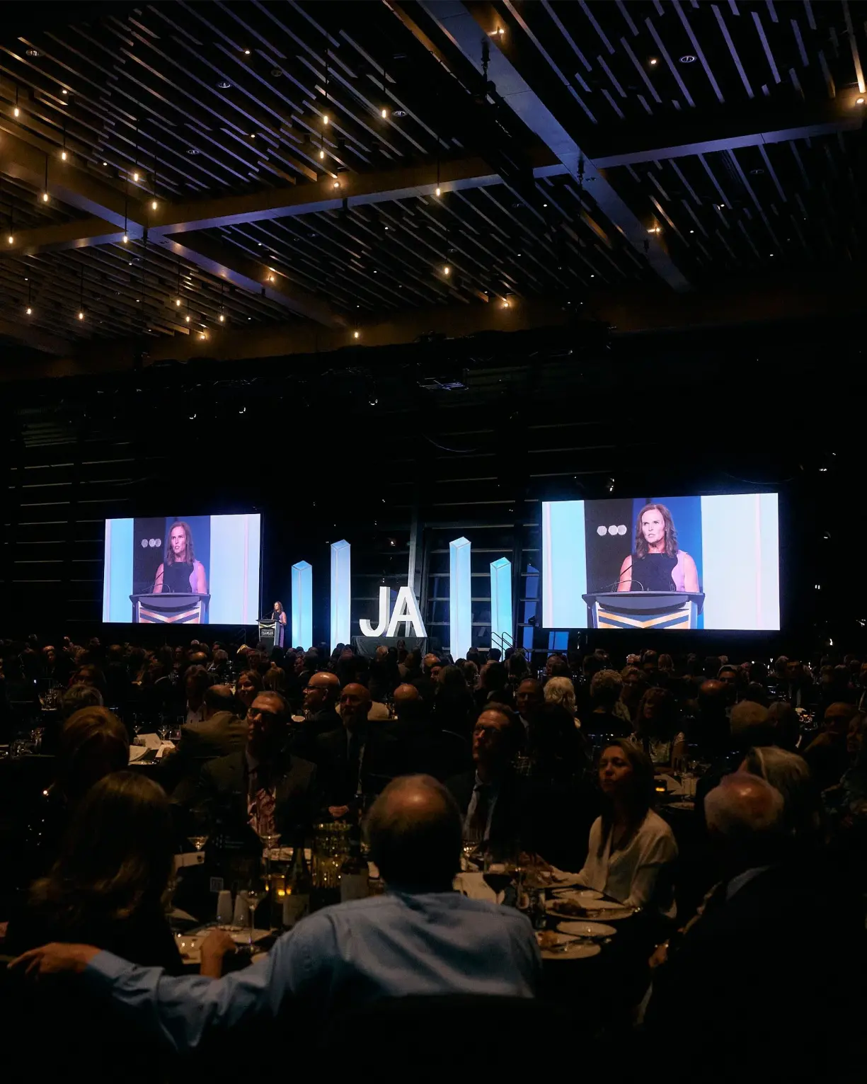Speaker on large screen at JA gala with audience seated at tables