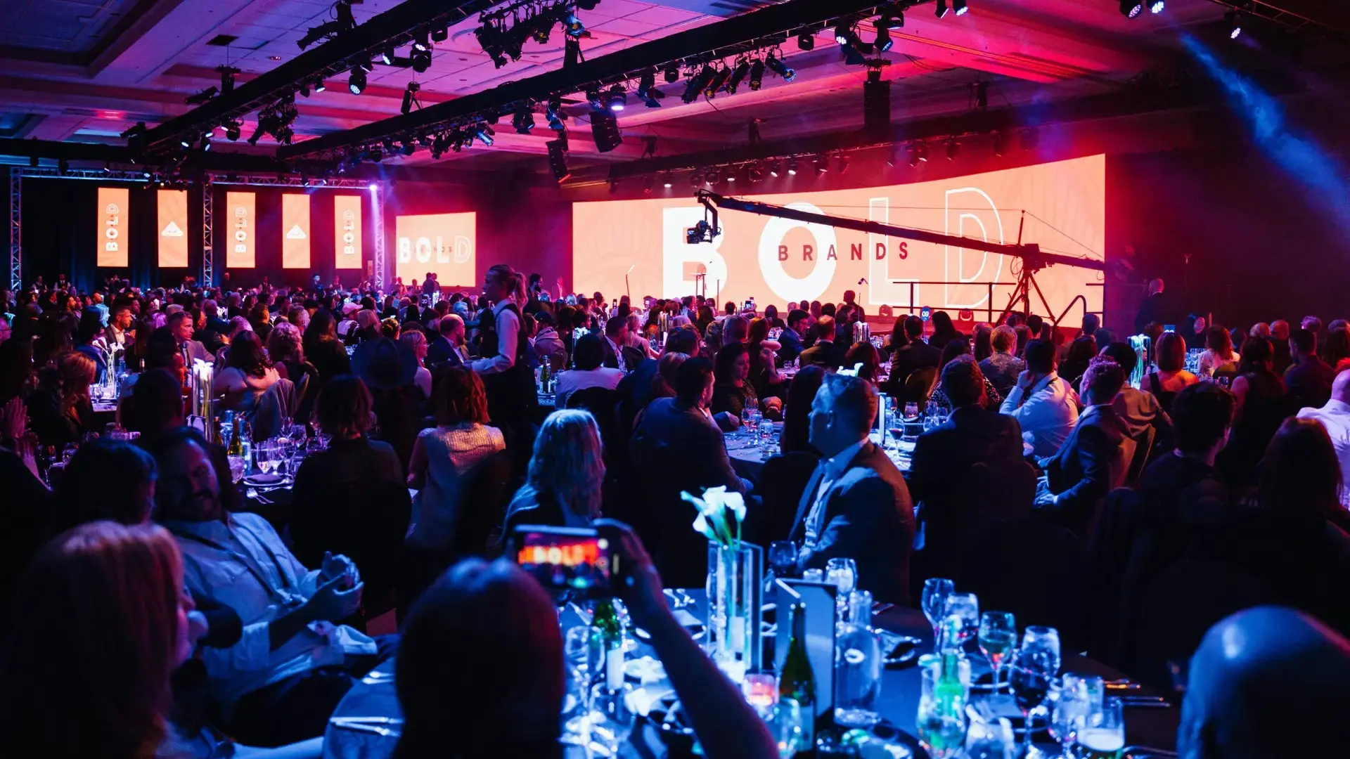 Crowd at a gala event with a large screen displaying BOLD BRANDS logo under stage lighting.