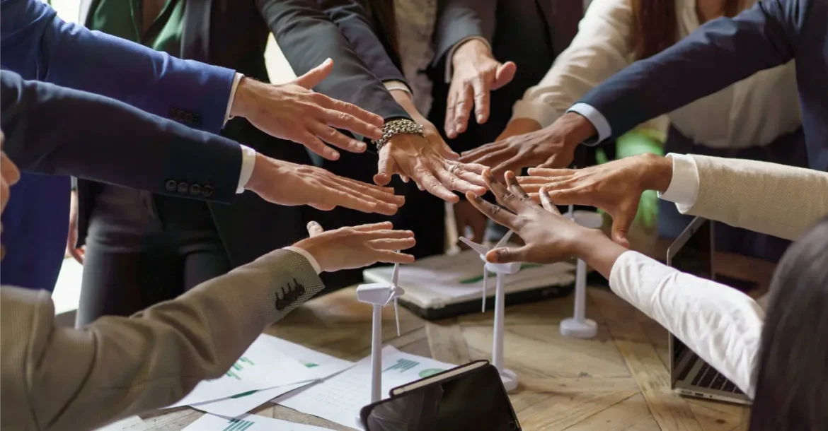 Diverse business team with hands stacked on table with wind turbine models, laptops and charts.