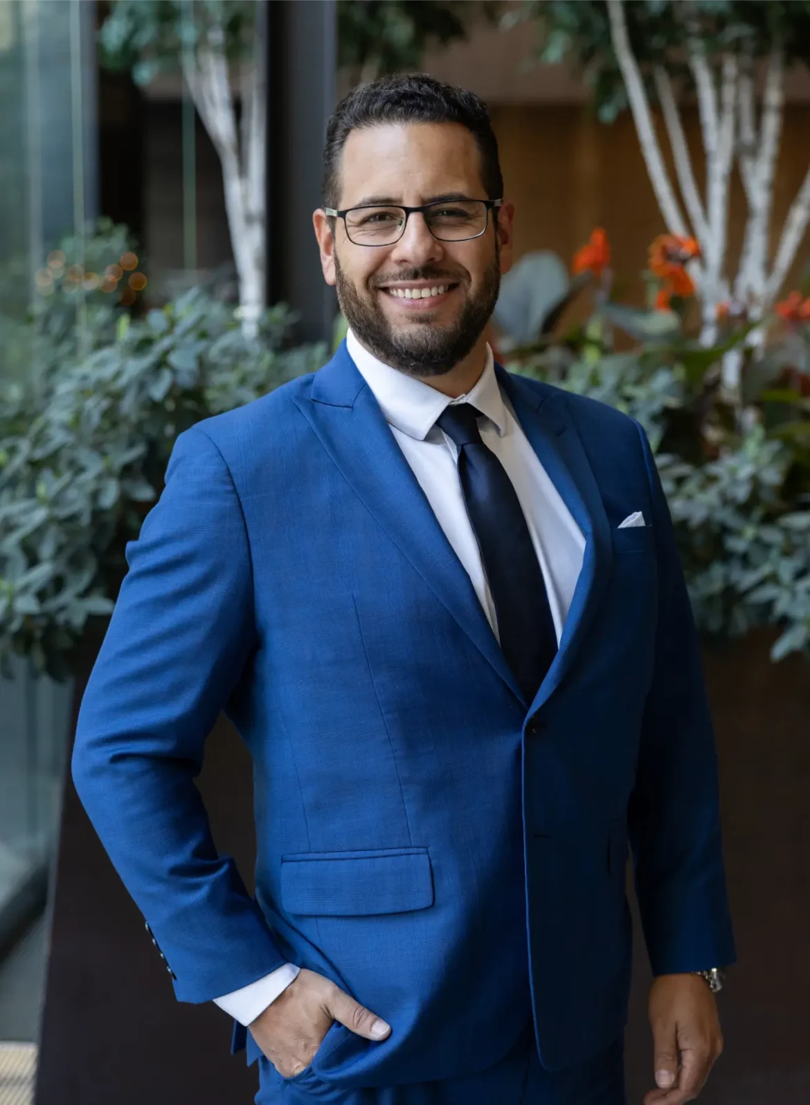 Man in blue suit with tie and glasses smiles, hand in pocket.
