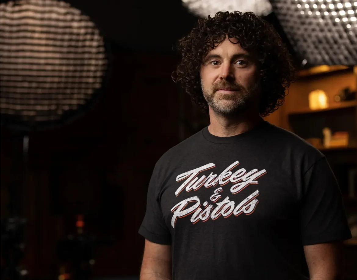 Man with curly hair wearing a black t-shirt with "Turkey & Pistols" printed on it