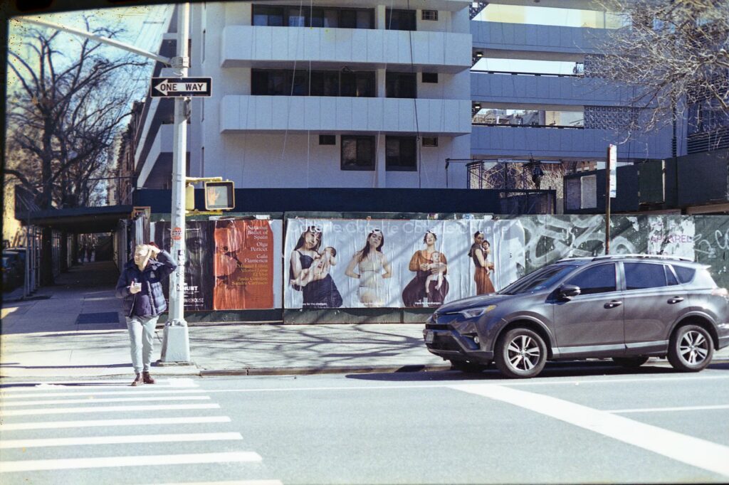 A pedestrian crossing the street in NYC amidst urban buildings and a parked vehicle.