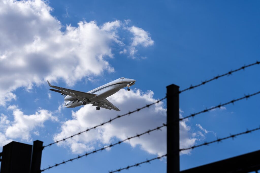 Jet approaching for landing, silhouetted against a bright blue sky with clouds and barbed wire in foreground.