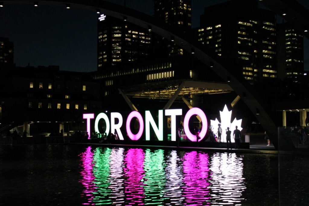 Illuminated Toronto sign reflecting over water at Nathan Phillips Square during night.