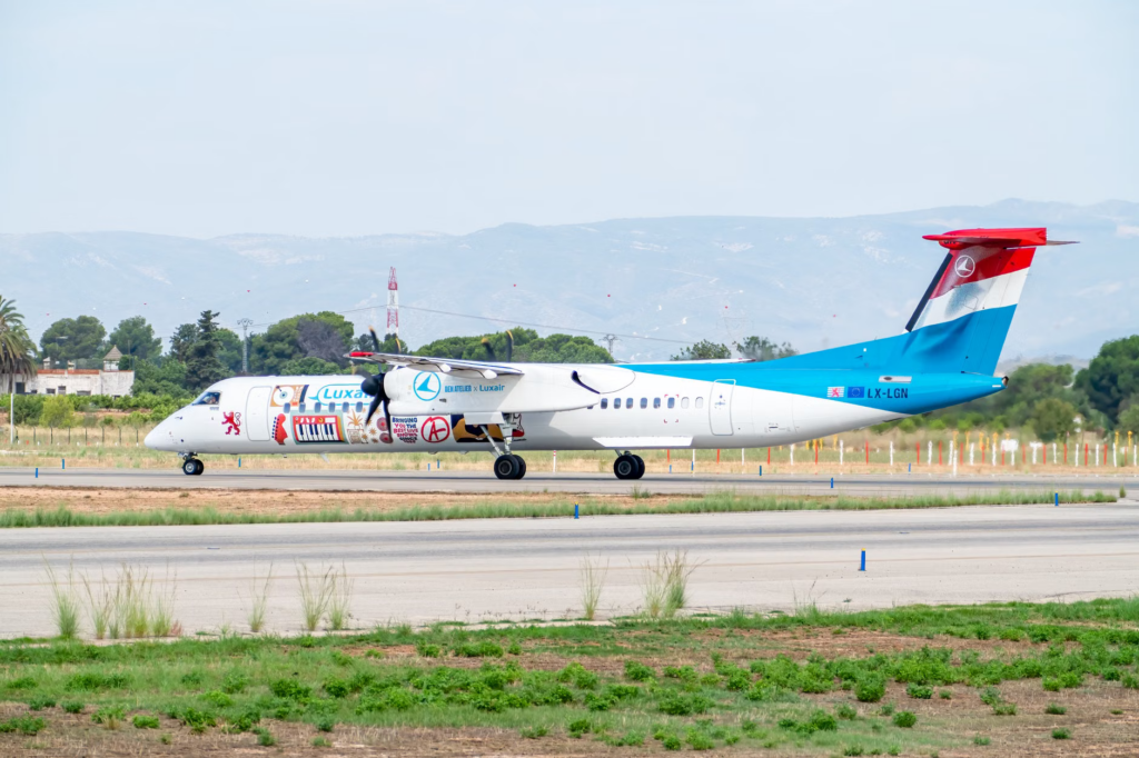 Colorful Luxair Q400 aircraft taxiing on a runway in Manises, Spain with a scenic backdrop.