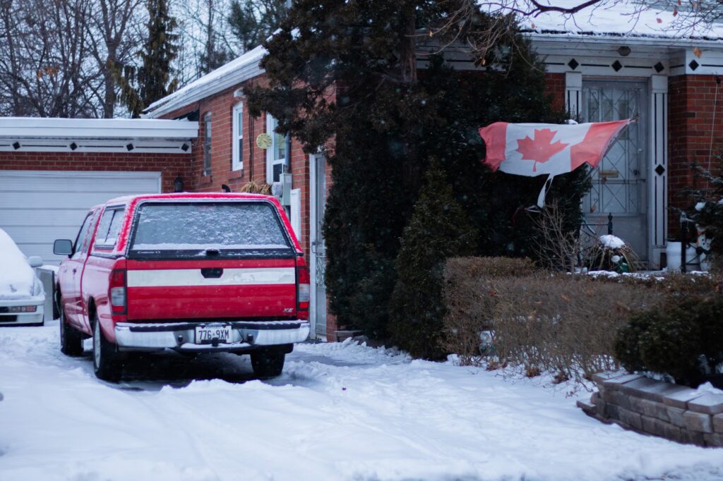 A red truck parked in a snowy Toronto front yard with a Canadian flag waving by the brick house.